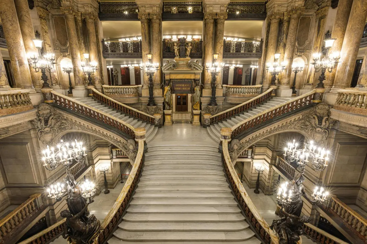 Grand escalier dans un bâtiment somptueux, où l'on aperçoit des rampes décoratives, des lustres et des statues.