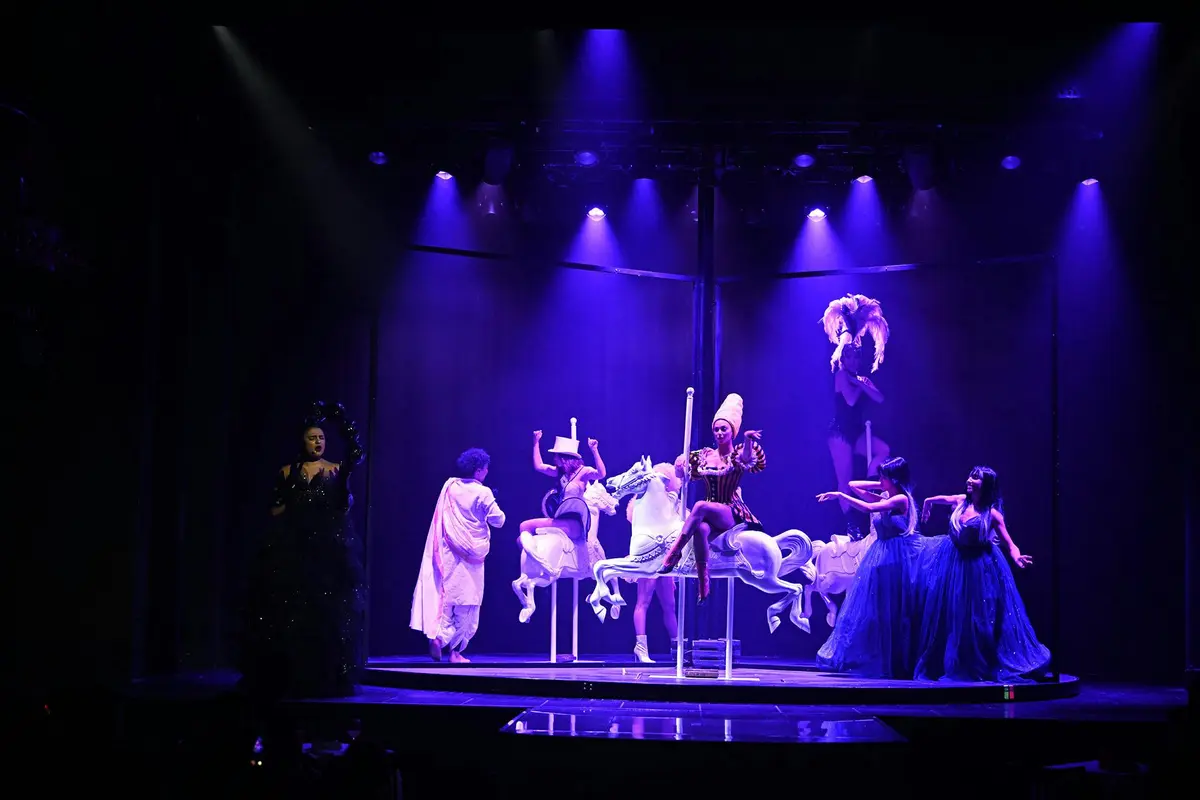 Des artistes sur une scène de carrousel, éclairée par des lumières violettes, avec une personne en costume sombre et d'autres en costume autour.