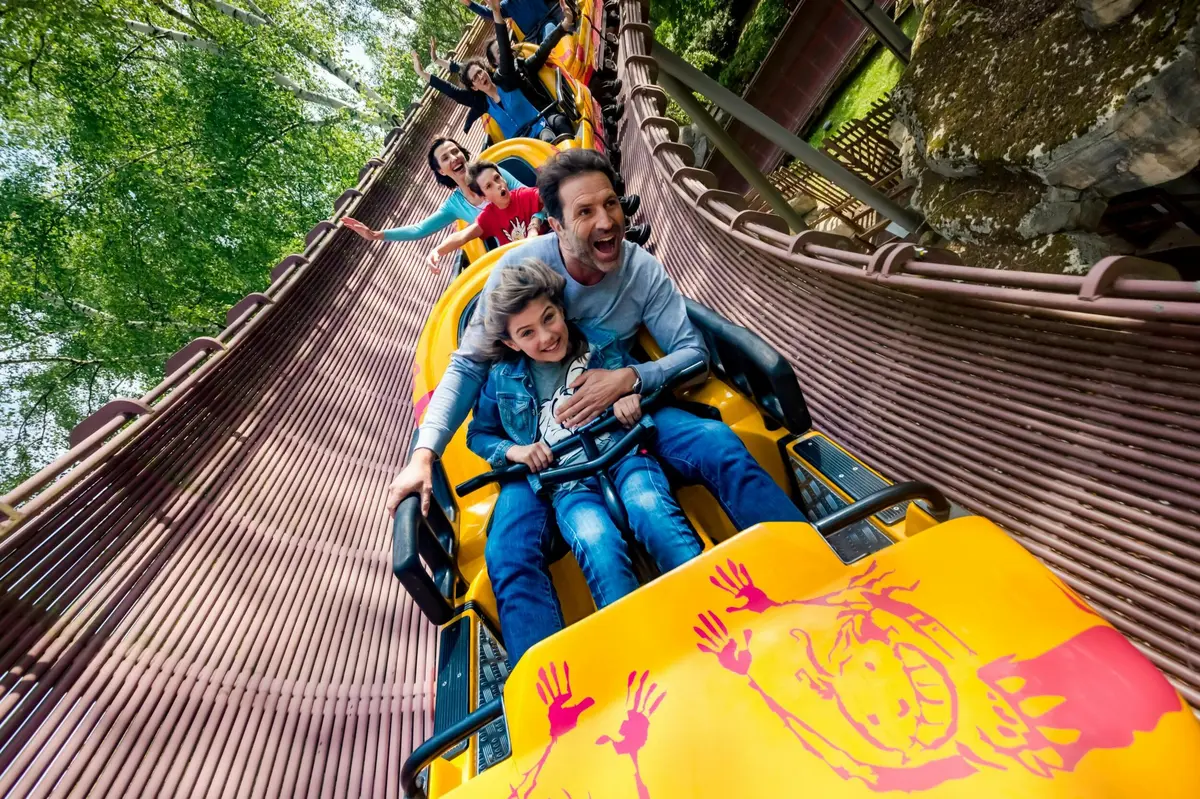 Des personnes à bord d'un grand huit jaune sur une piste sinueuse en bois, souriantes et visiblement enthousiastes.