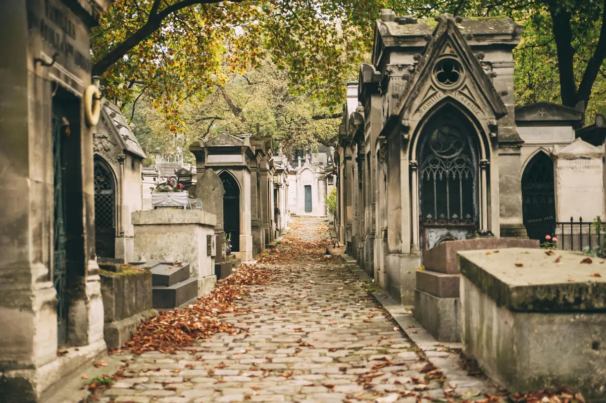 Chemin pavé étroit bordé de tombes ornées et de feuilles d'automne, sous des arbres au feuillage vert et jaune.