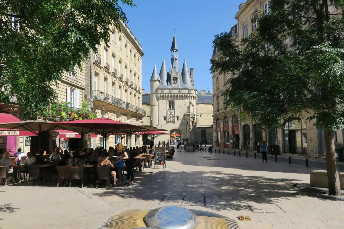 Outdoor caf&eacute; seating under red umbrellas, near historic building with turrets on a sunlit street. Trees on either side.