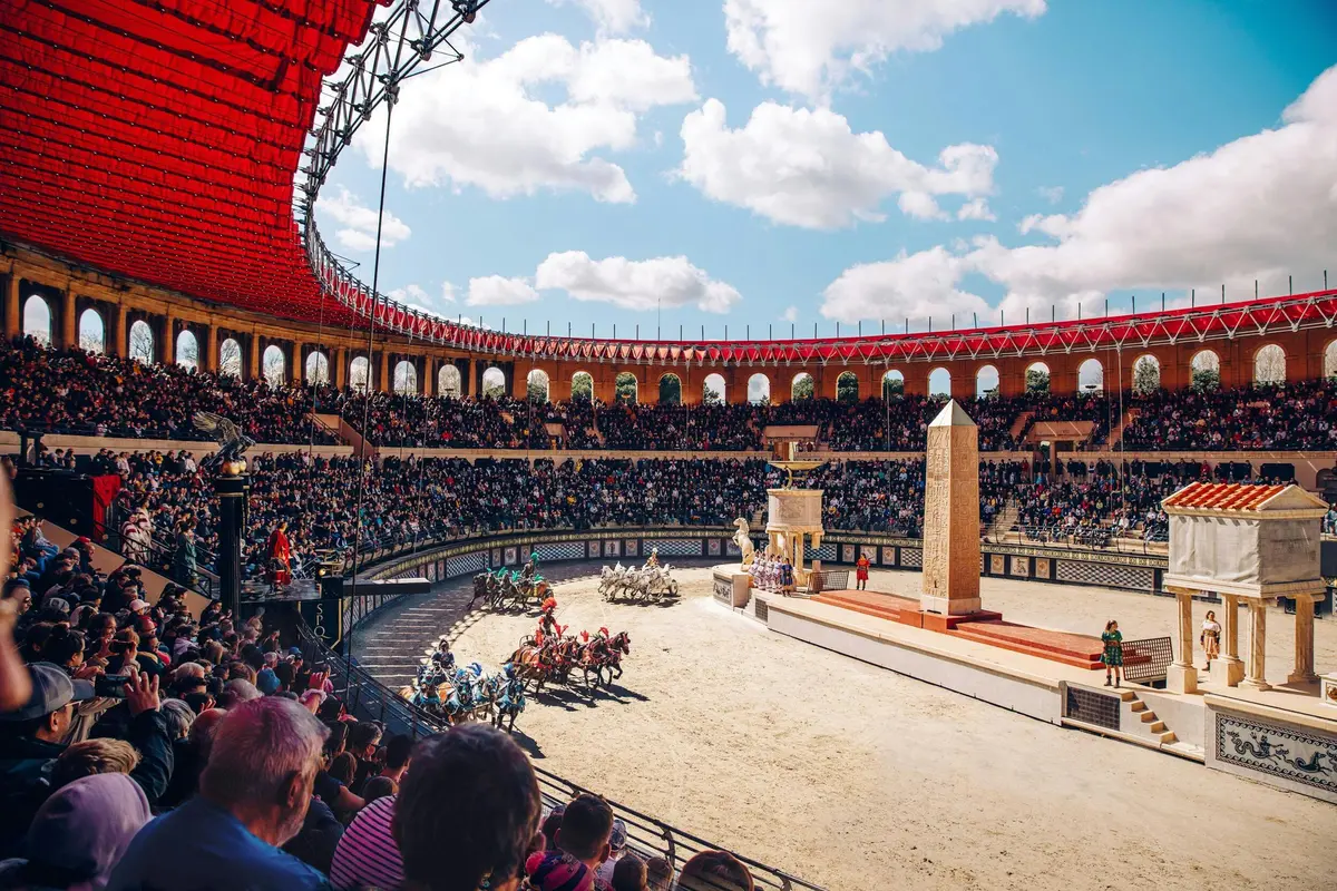 Une course de chars tirés par des chevaux dans une grande arène avec des sièges en gradins remplie de spectateurs sous un ciel partiellement couvert.