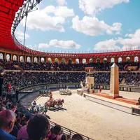 Une course de chars tirés par des chevaux dans une grande arène avec des sièges en gradins remplie de spectateurs sous un ciel partiellement couvert. DR