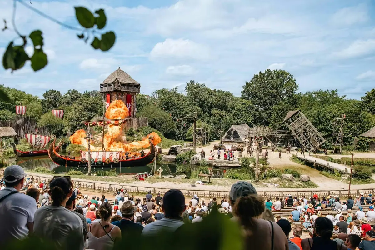 Spectacle de théâtre en plein air mettant en scène une scène viking avec un bateau en feu, des comédiens et le public, sous un ciel bleu.
