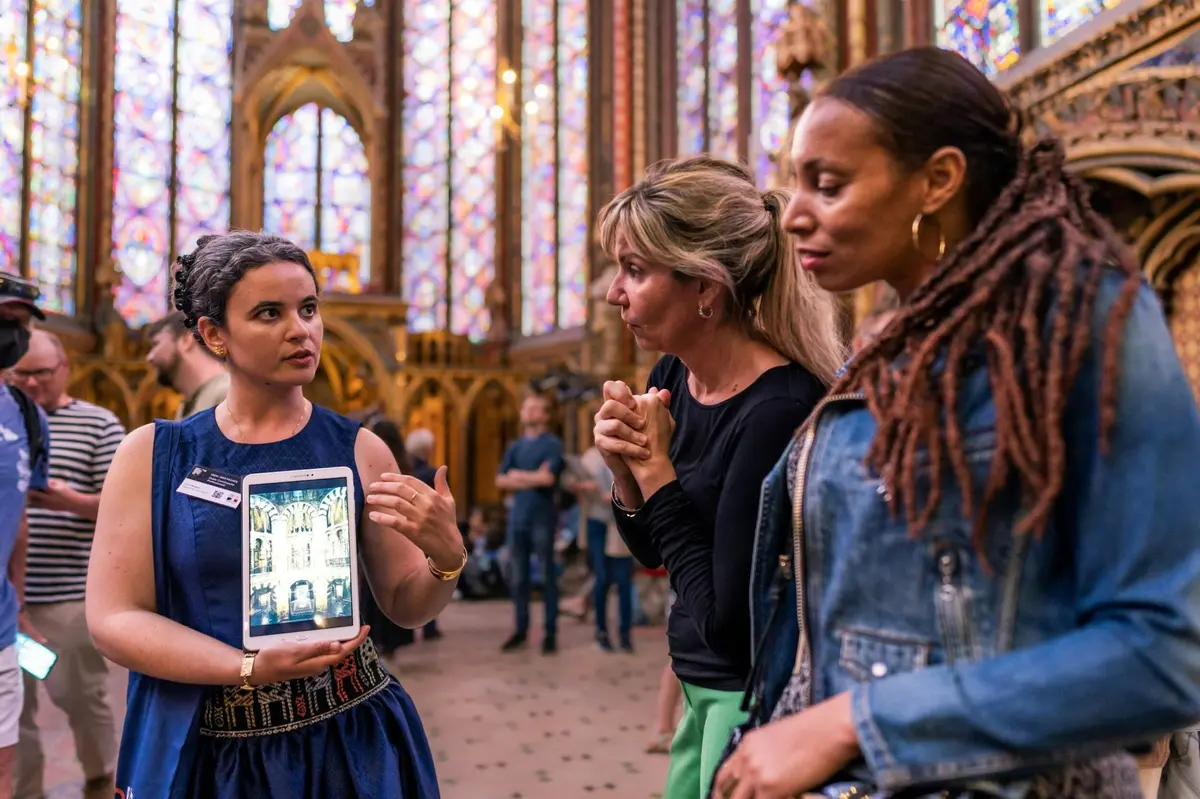 Une femme en robe bleue montre une image sur une tablette à deux autres personnes dans un bâtiment de style gothique avec des vitraux.