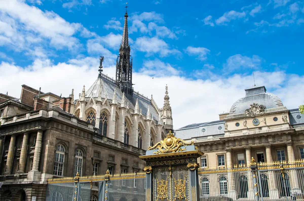 Bâtiment de style gothique orné d'une haute flèche, de sculptures en pierre complexes et de portes décoratives sous un ciel bleu parsemé de nuages.