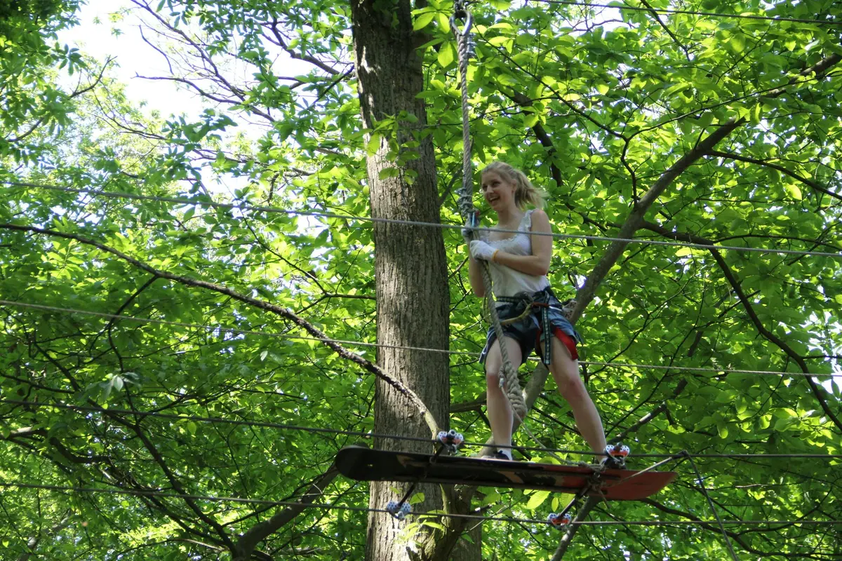 Une personne se tient en équilibre sur une planche suspendue au milieu des arbres, sourit, porte un harnais et s'accroche à des cordes dans une zone forestière.
