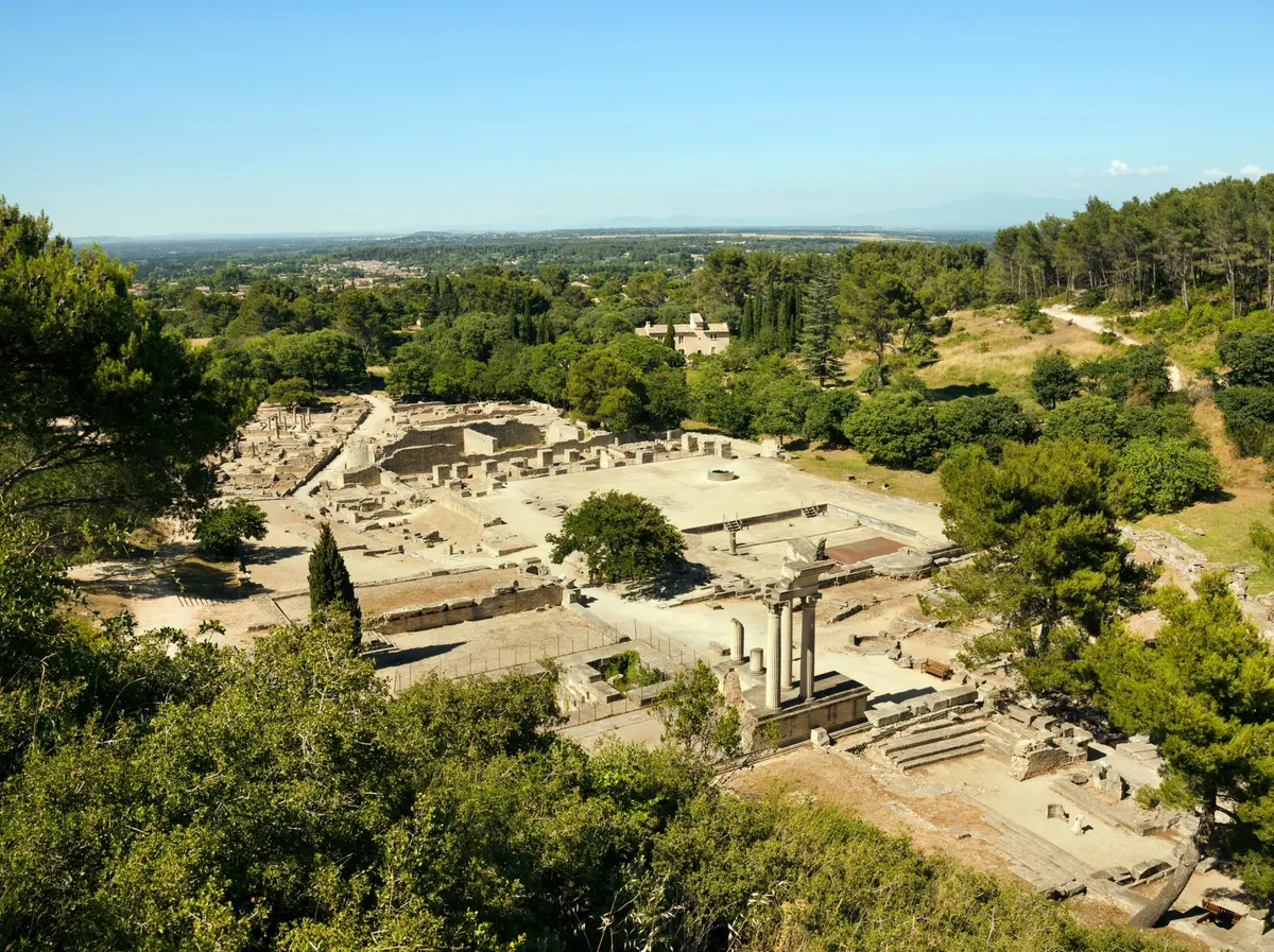 Des ruines anciennes avec des colonnes debout entourées d'arbres, avec une vue lointaine sur la campagne sous un ciel bleu clair.