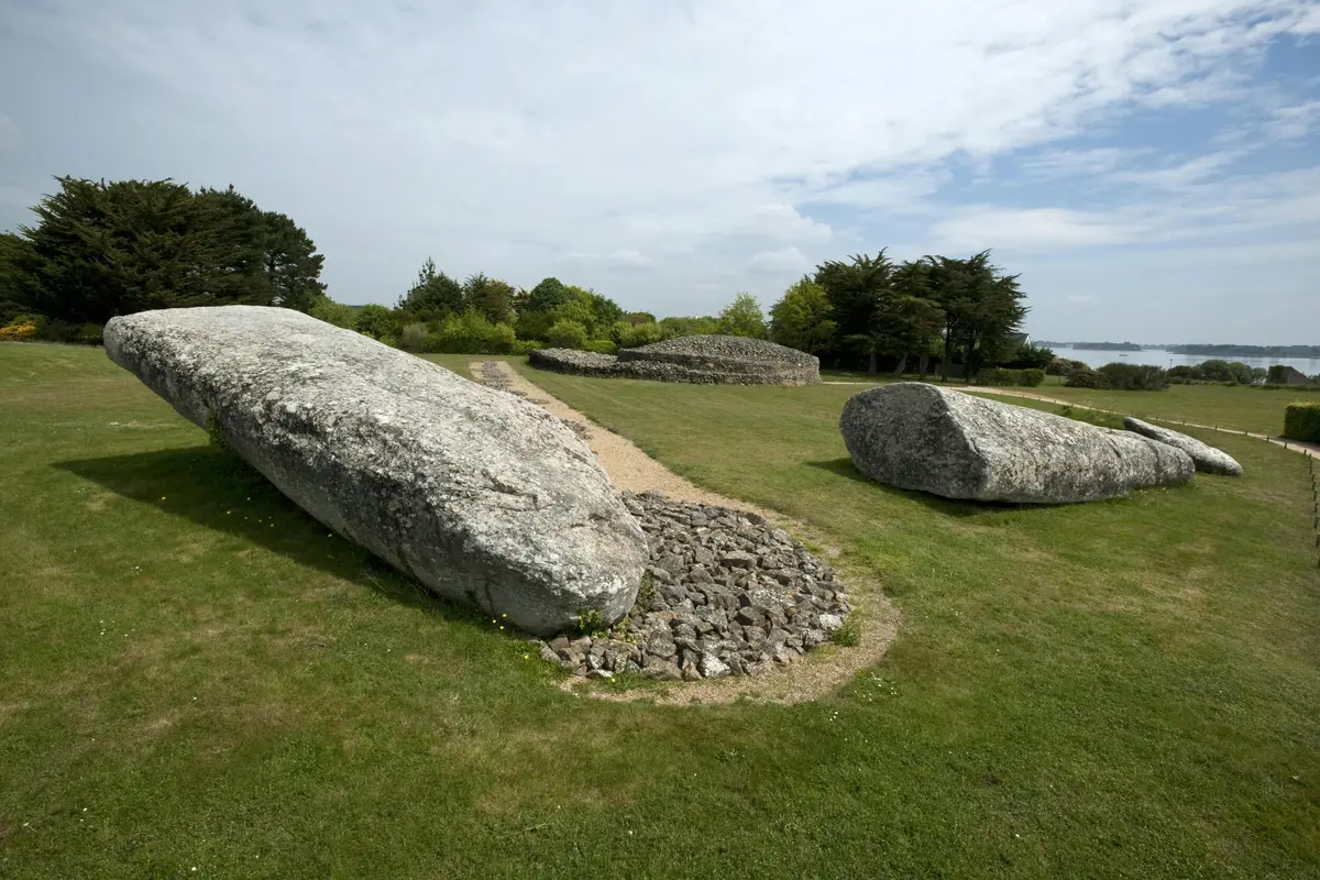 Grandes pierres anciennes entourées d'herbe et d'un chemin de gravier, avec des arbres et un ciel nuageux en arrière-plan.