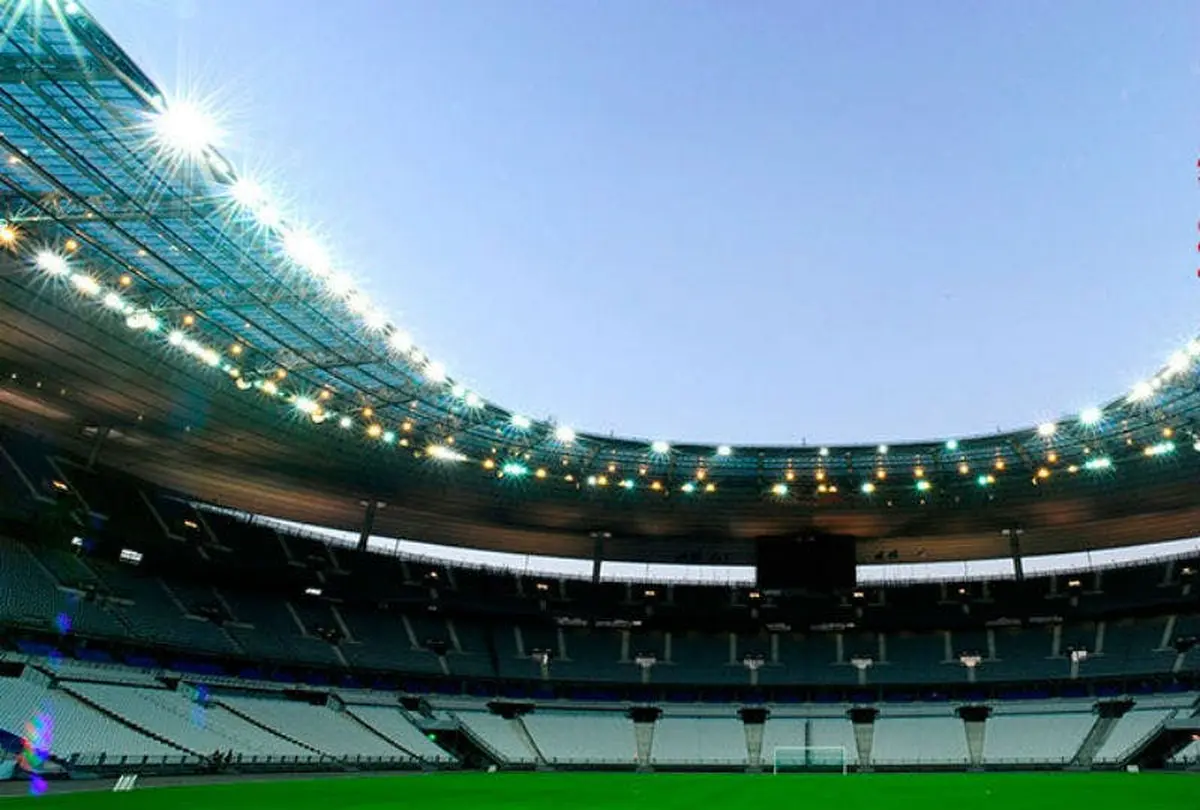 Un grand stade vide, avec des sièges en gradins et des lumières vives, montrant un terrain vert et un ciel clair au-dessus.