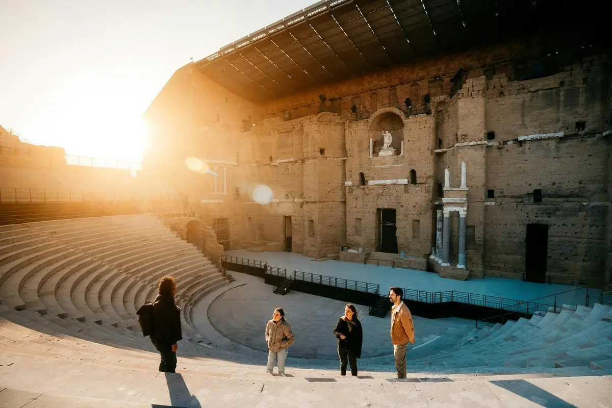 Un groupe de quatre personnes se tient dans un amphithéâtre antique, dont la grande scène en pierre est baignée par la lumière du soleil.