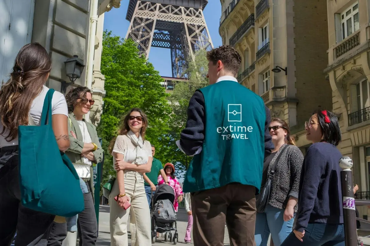 Un groupe de personnes, dont certaines portent des sacs, discutent près d'un immeuble, avec la Tour Eiffel en arrière-plan.