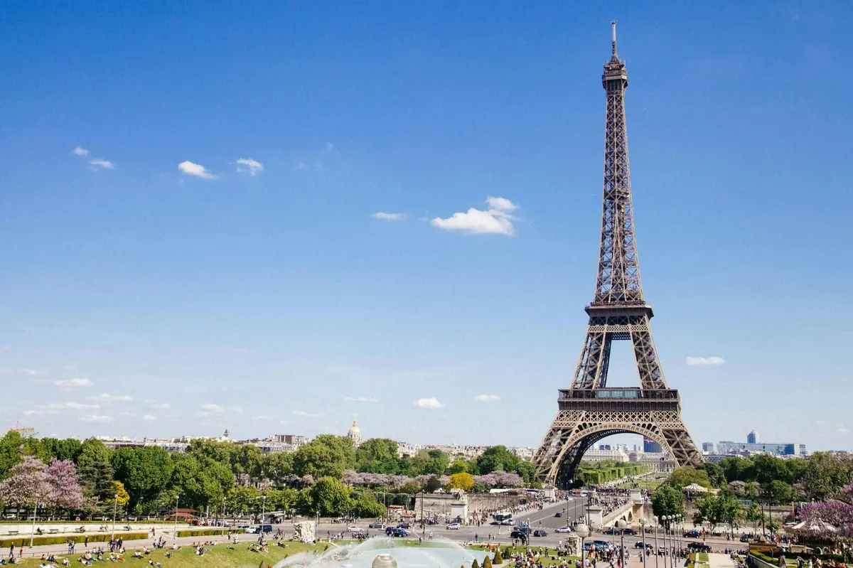 La Tour Eiffel dans un parc sous un ciel bleu clair, avec la verdure environnante et les personnes visibles au premier plan.