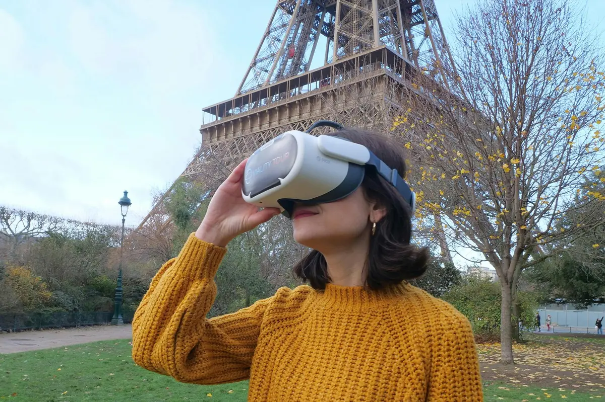A person in a yellow sweater uses a virtual reality headset near the Eiffel Tower in a park setting on a clear day.