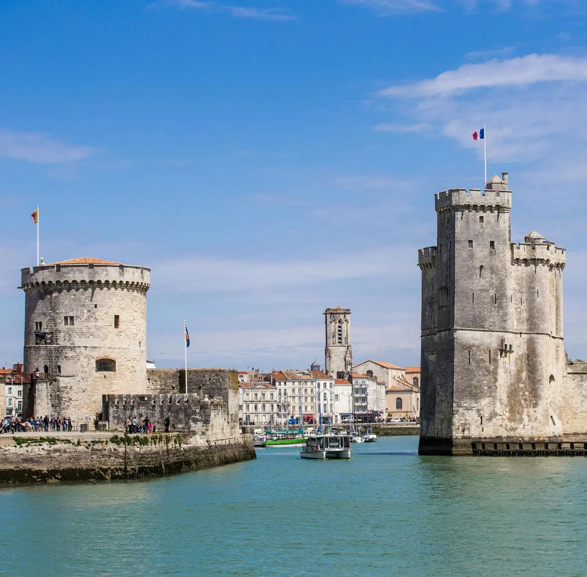Deux tours en pierre avec des drapeaux s'élèvent à côté d'un canal sous un ciel bleu ; des personnes et des bateaux sont visibles près des bâtiments historiques.