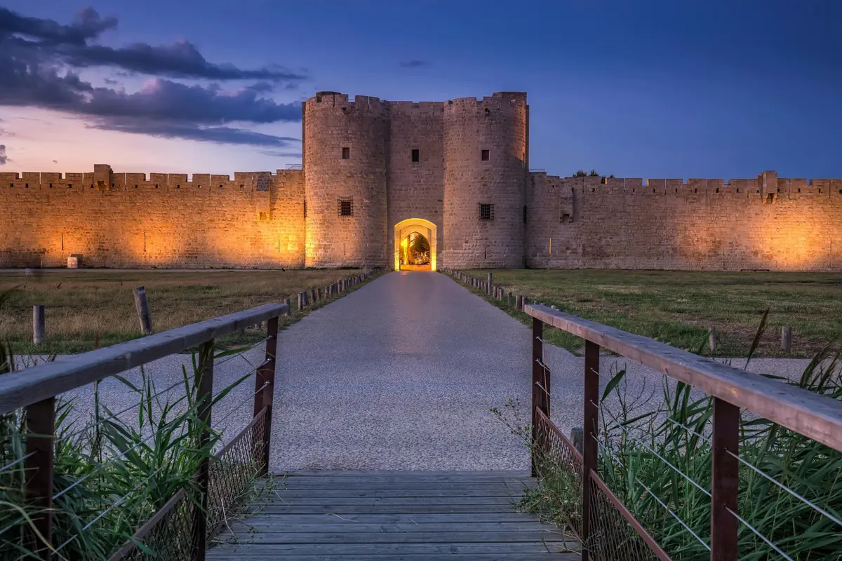 Château de pierre aux murs illuminés à la tombée de la nuit, vu depuis un pont de bois et le chemin menant à l'entrée.