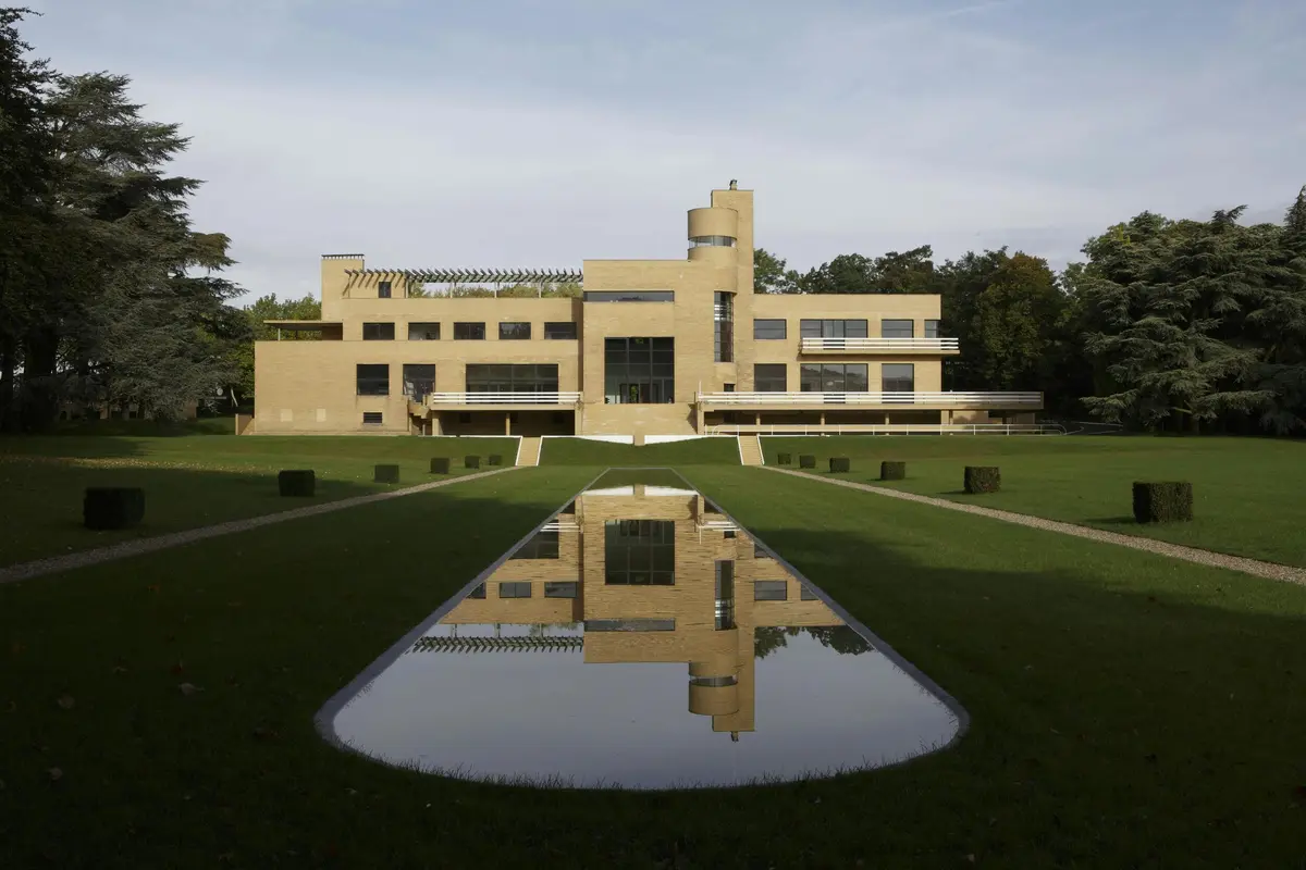 Un grand bâtiment moderne en briques au design symétrique se reflète dans une piscine rectangulaire située dans un jardin bien entretenu. Des arbres se trouvent à l'arrière-plan.