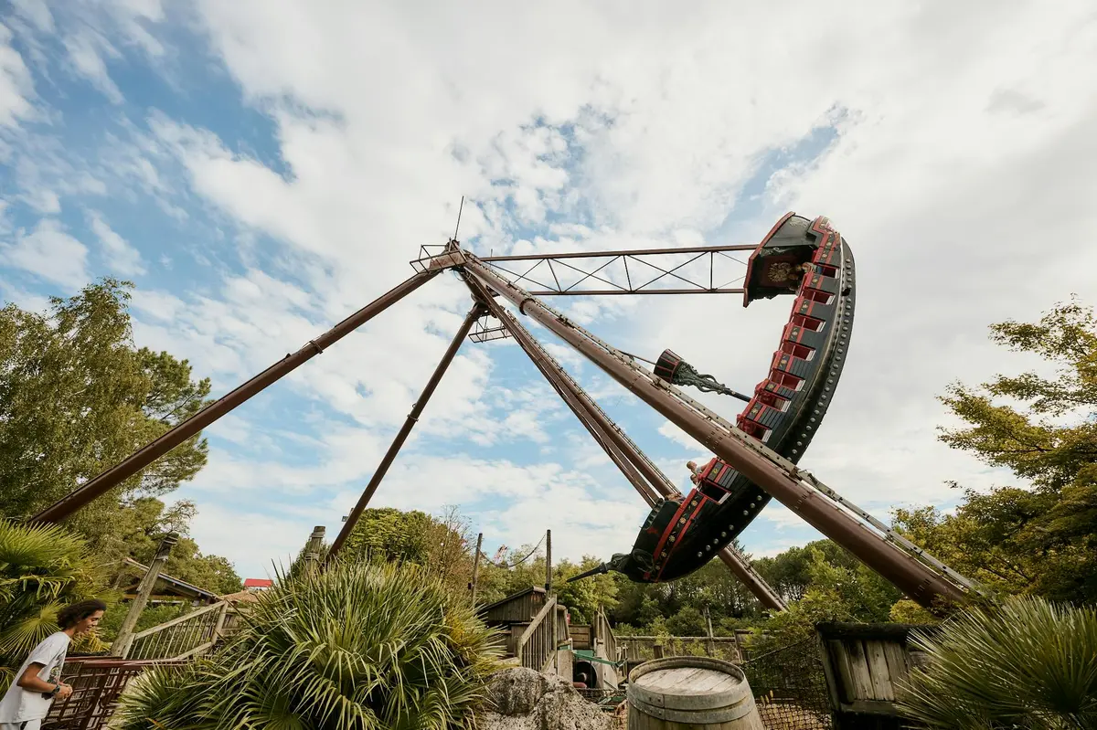Un grand bateau de pirates se balance dans les airs dans un parc d'attractions, entouré d'arbres et d'un ciel partiellement nuageux.