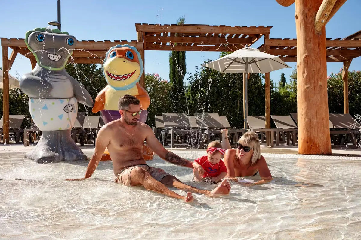 Un homme, une femme et un jeune enfant jouent dans une zone d'eau peu profonde, près d'une statue de requin aux couleurs vives et de transats installés sous des parasols.