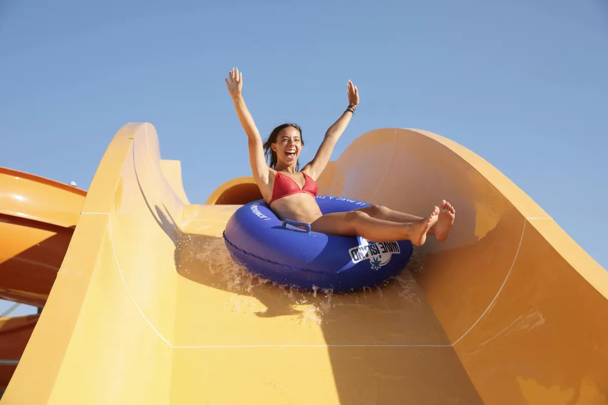 Une femme en bikini rouge se promène sur une chambre à air bleue sur un toboggan aquatique jaune vif sous un ciel bleu clair.