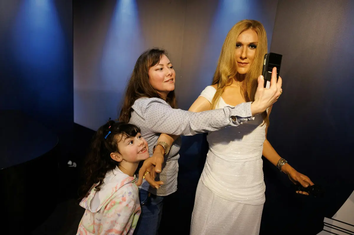 Une femme et un enfant posent à côté d'une statue de cire représentant une femme vêtue de blanc, tandis que celle-ci tient un téléphone pour prendre un selfie.